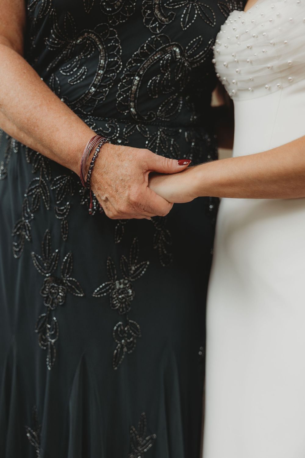 Bride in white gown holds hands with woman in black beaded dress, sharing a tender moment at a wedding.