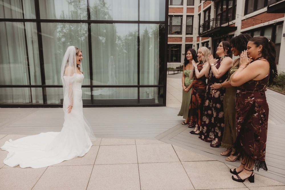 Bride in white gown and veil stands before emotional bridesmaids in colorful dresses during outdoor first look.