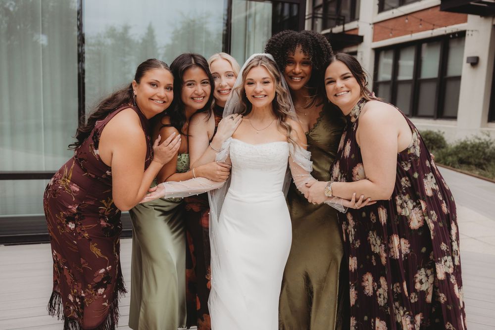Bride in white gown poses with five bridesmaids in mixed green and floral dresses, smiling outdoors.
