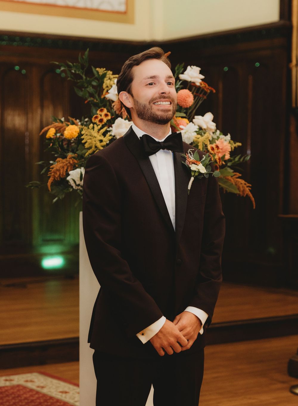 Smiling groom in black tuxedo with bow tie stands at wedding altar with colorful floral arrangements behind him.