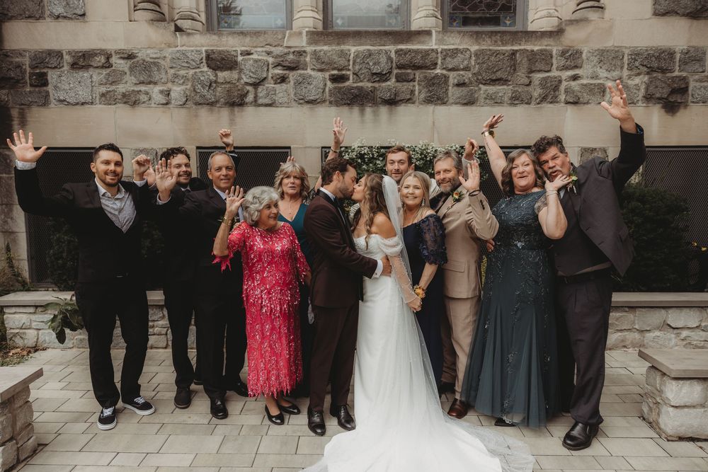 Wedding party celebrates with newlyweds kissing outdoors at a stone venue, guests cheering with raised arms.