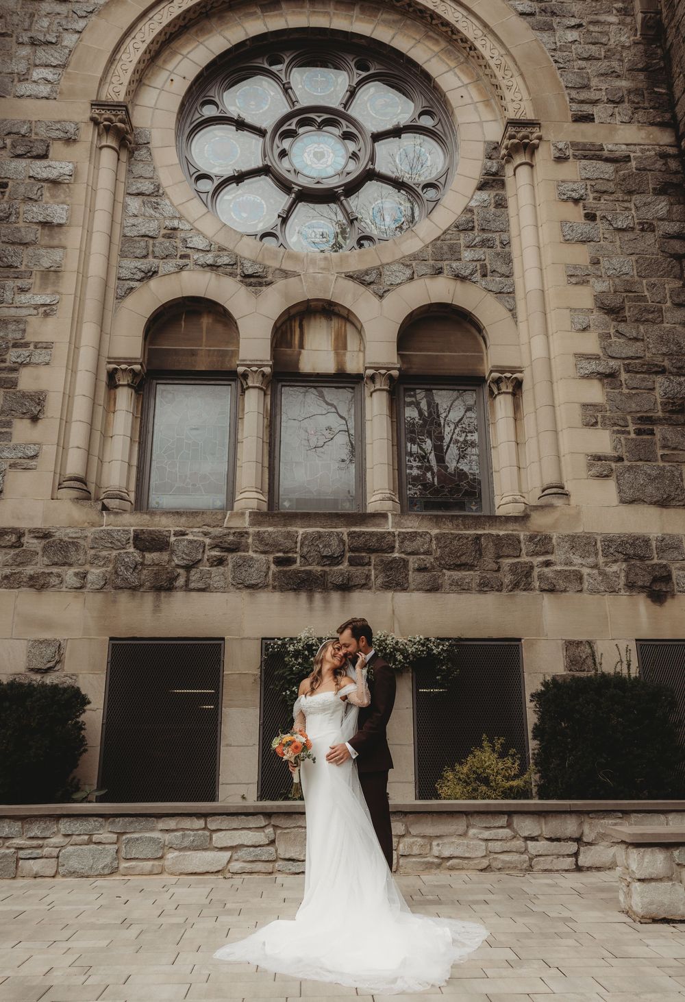 Bride and groom kissing in front of a stunning stone church with a rose window on their wedding day.