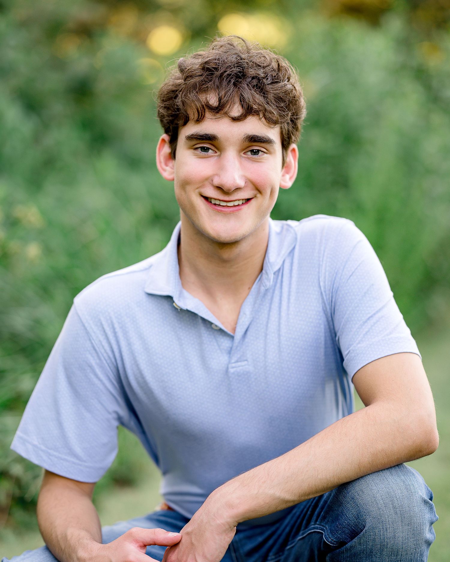 Smiling young man with curly hair wearing a light blue polo shirt, seated outdoors with green foliage background.