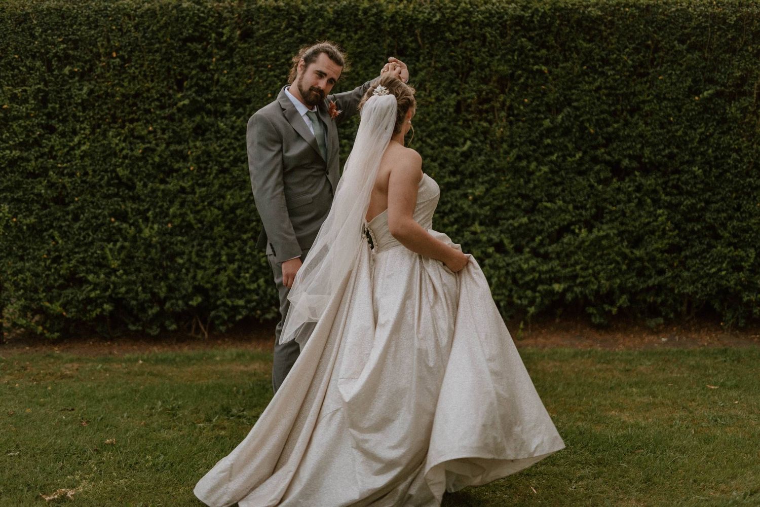 A bride in a flowing white gown and veil twirls outdoors while her groom in a gray suit watches lovingly.