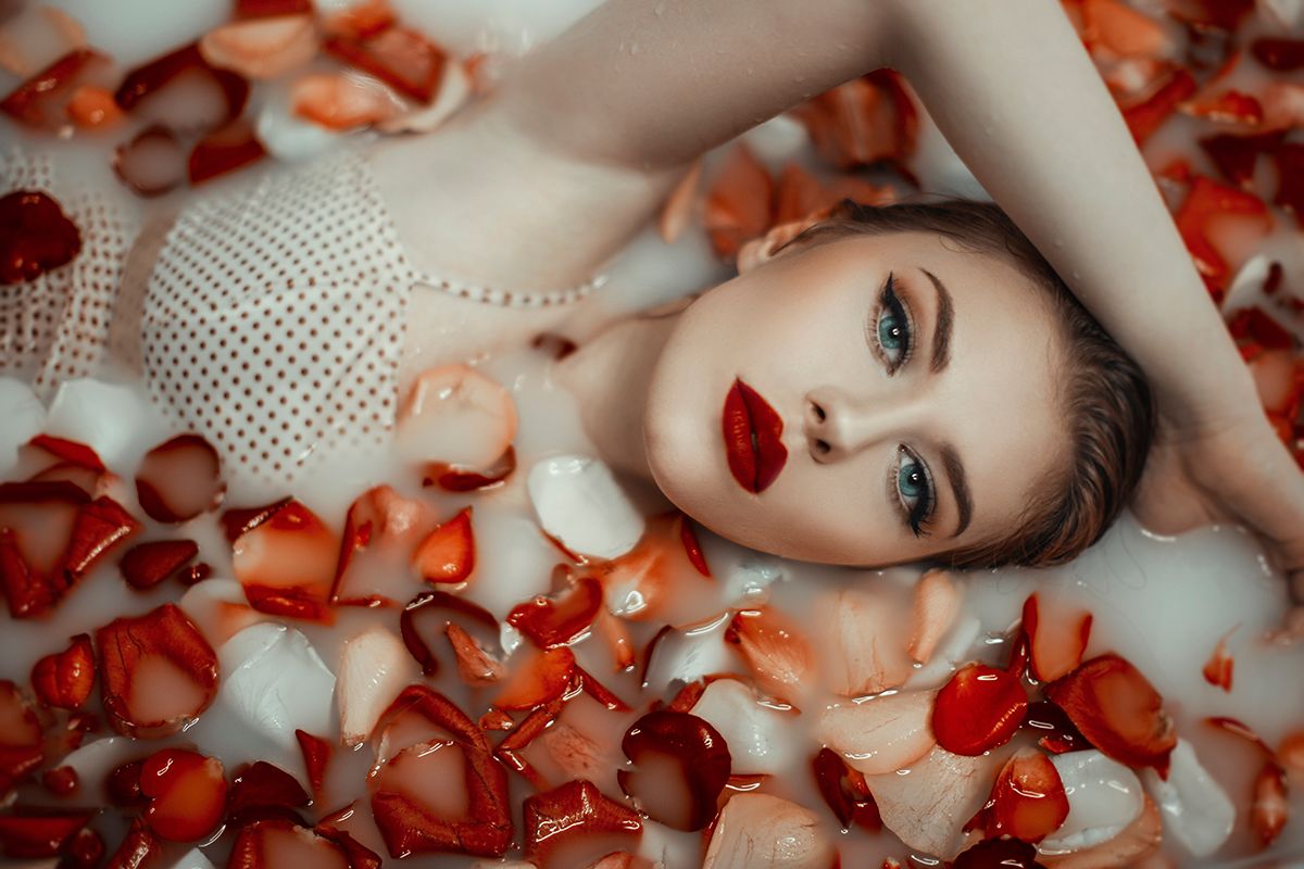 Woman with red lipstick lying in a milk bath filled with red rose petals