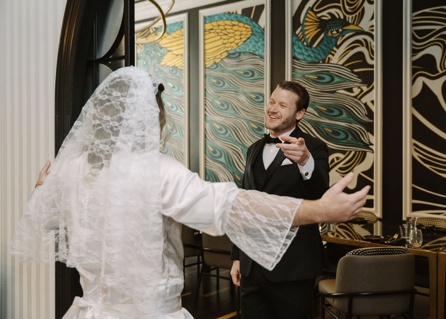Groom in black tuxedo reacts with joy as bride in lace veil approaches during wedding first look ceremony.
