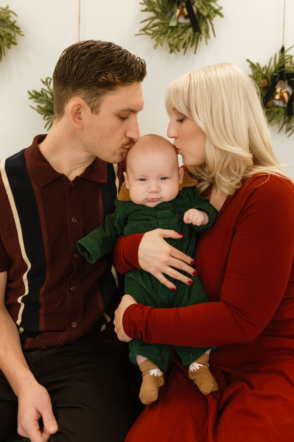 A couple in festive attire kisses their baby dressed in green during a Christmas family portrait session.