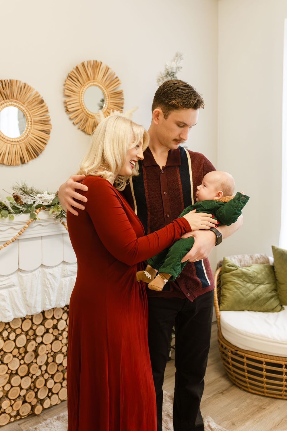 A young family in festive attire poses together, with the father holding a baby in green near a decorated fireplace.
