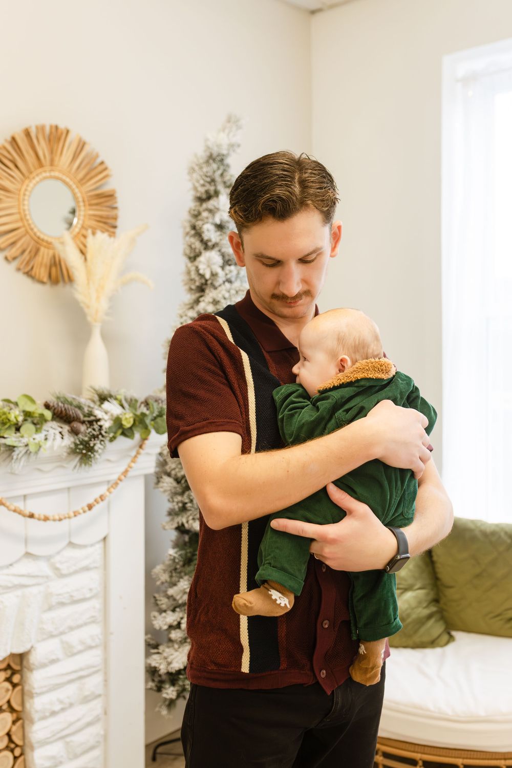 A young father holding his infant baby dressed in green near a decorated Christmas tree indoors.