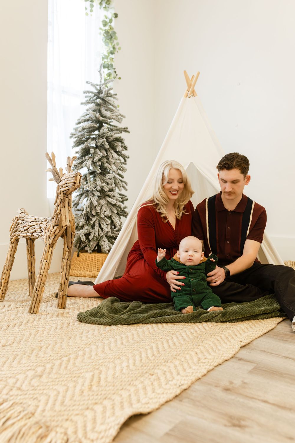 Family Christmas photo with parents and baby sitting on rug near teepee, reindeer decor, and flocked tree.