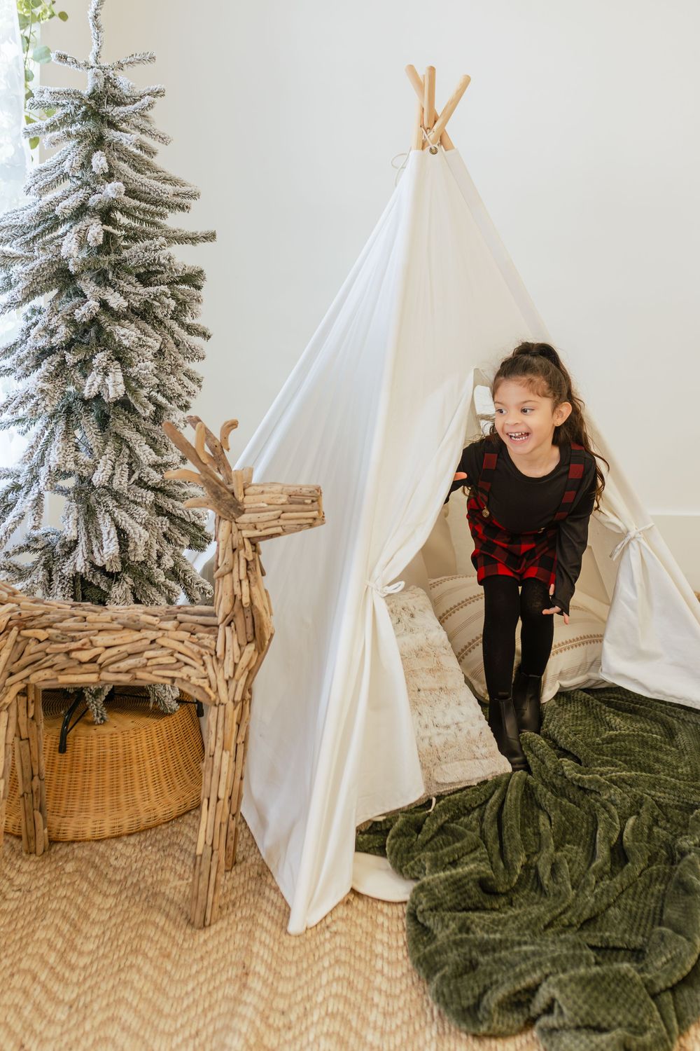 A happy young girl in plaid pajamas plays beside a white teepee tent, with a flocked Christmas tree and wooden reindeer decor.