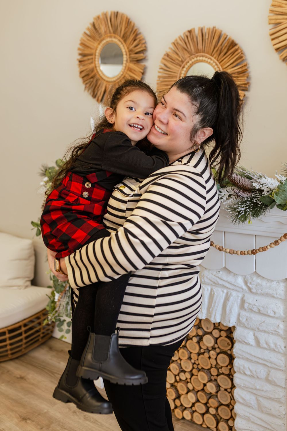 A mother in a striped shirt holds her smiling young daughter in a red plaid dress near a Christmas fireplace.