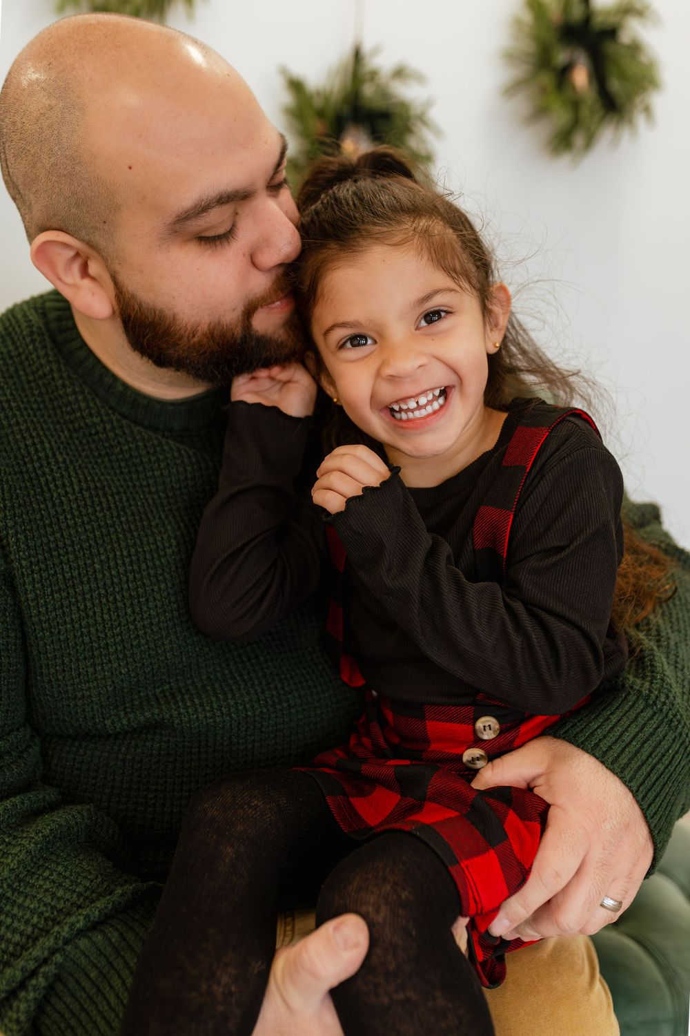 Father kissing daughter's cheek during Christmas photo session, girl smiling in red plaid dress.