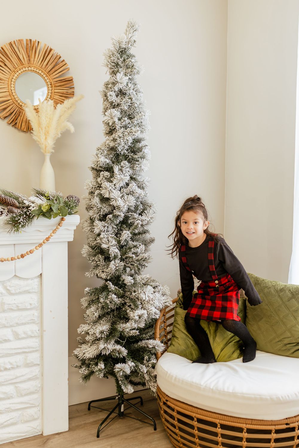 Young girl in red plaid dress smiling beside a slim flocked Christmas tree next to a white fireplace.