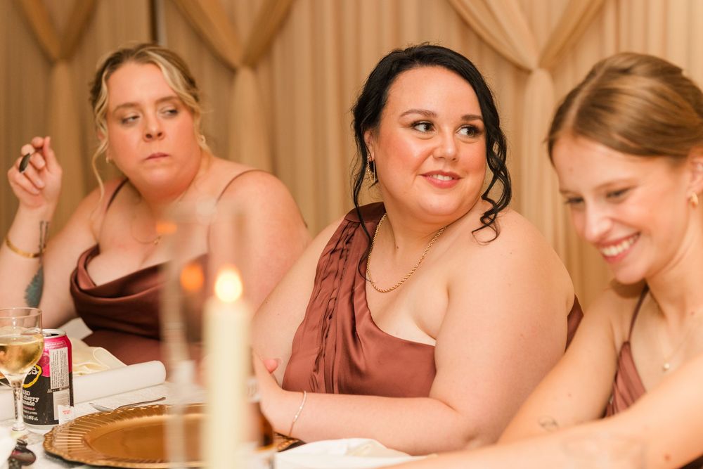 Three bridesmaids in satin dresses seated at a wedding reception table with candles and elegant decor.