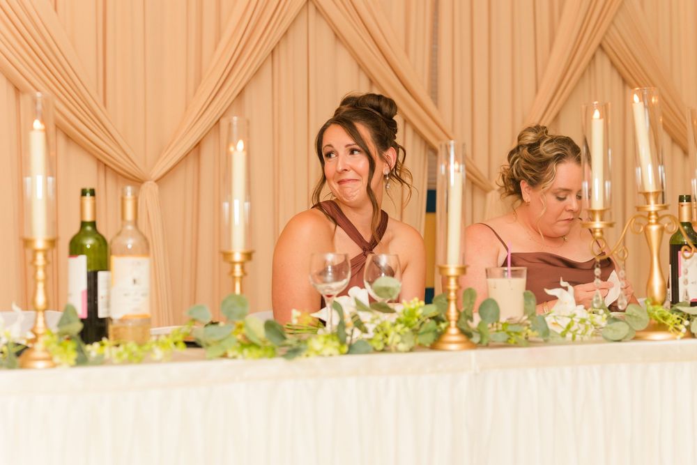 Two bridesmaids in burgundy dresses seated at a decorated head table with candles and greenery at a wedding reception.