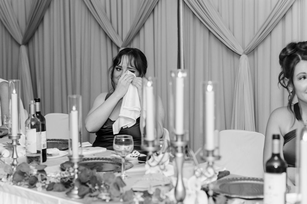 Emotional wedding guest dabbing tears with napkin at elegantly set reception table with candles and flowers.
