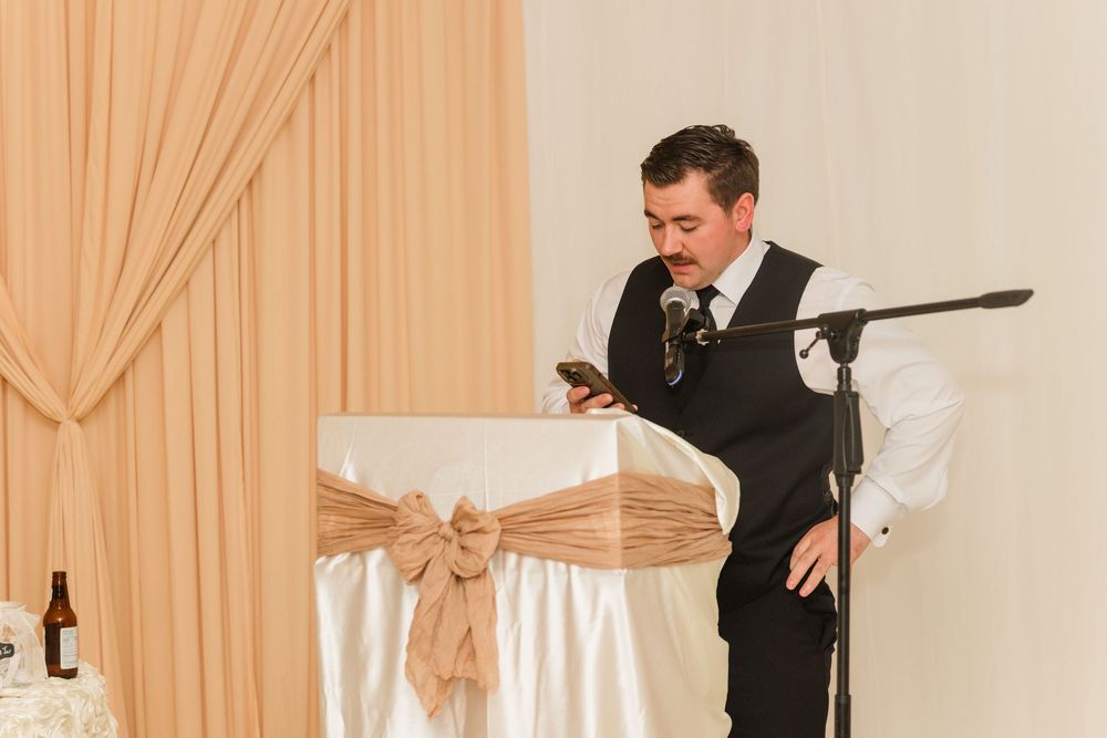 Man in vest reads from phone at wedding reception podium with elegant white and gold chair decor.