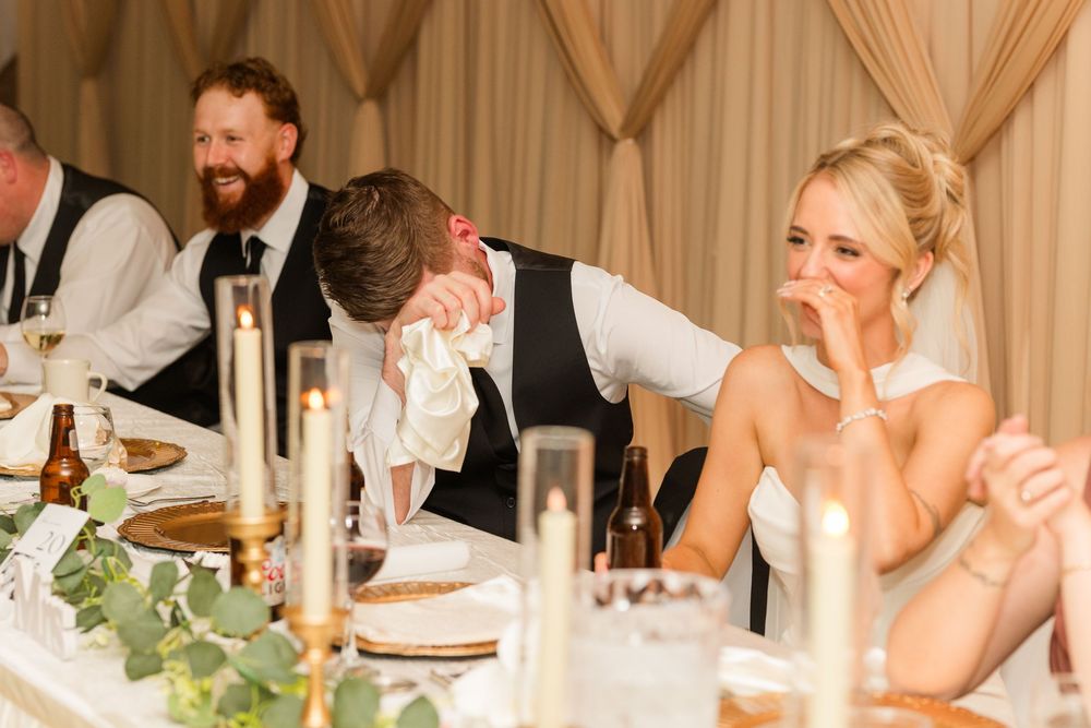 Bride and groom laughing during wedding reception speeches at elegantly decorated head table with candles.