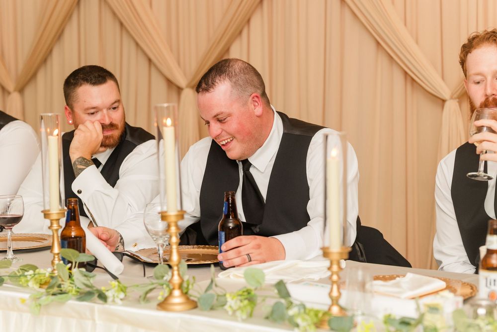 Groomsmen in black vests and white shirts laugh at a wedding reception table with gold candelabras.