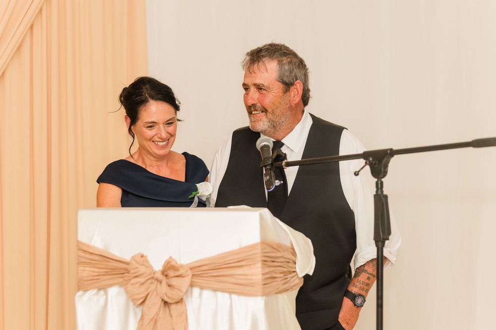 A man and woman share a laugh at a podium with a burlap bow during a wedding reception speech.