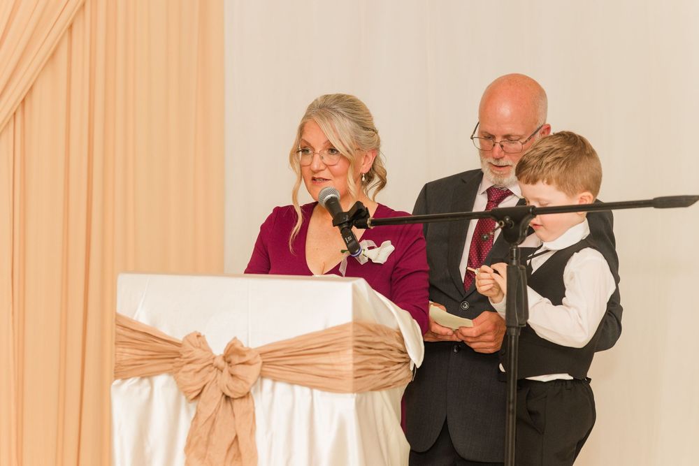 Woman speaking at podium with man holding young boy beside her at a formal wedding reception event.