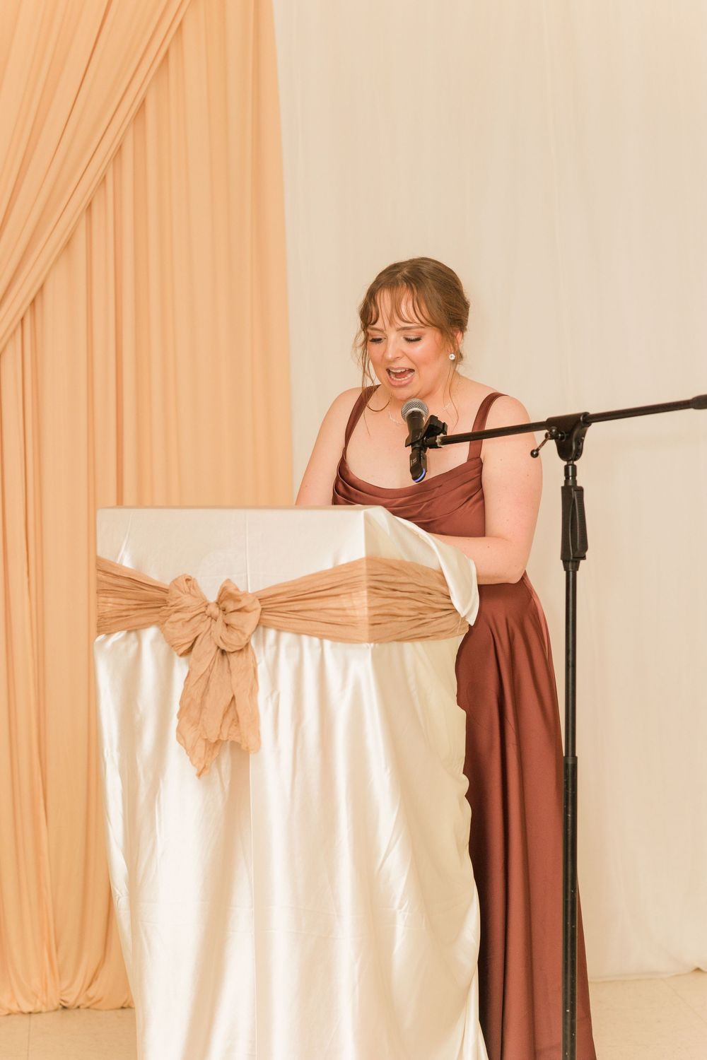 Woman in rust satin gown giving speech at wedding podium with microphone, elegant cream draping background.