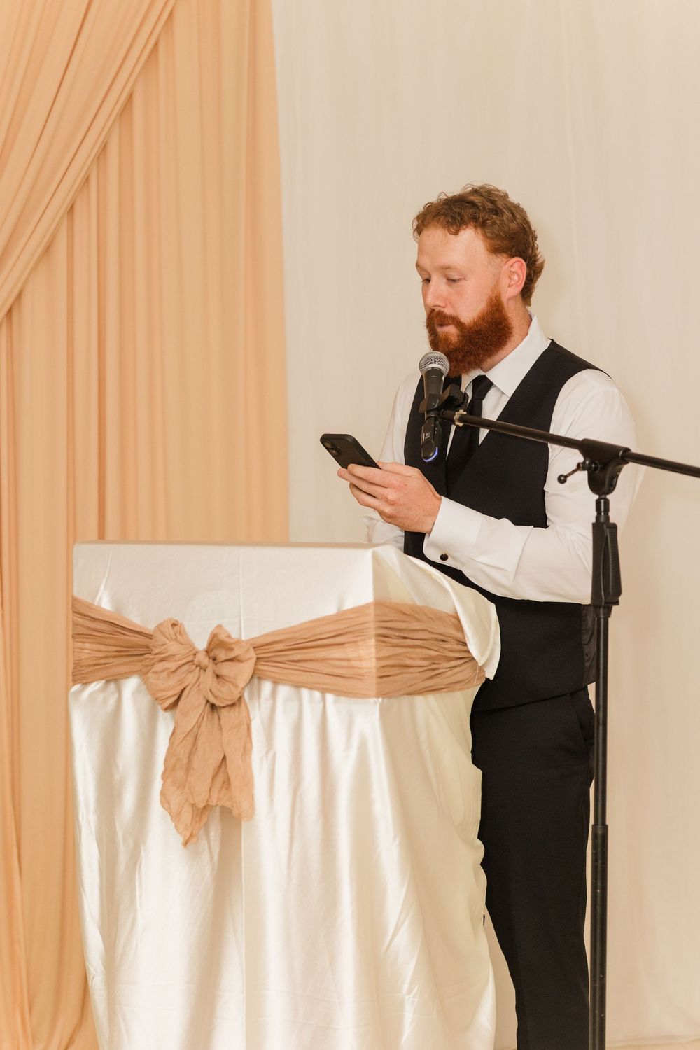 Bearded man in vest reads from smartphone at decorated podium with microphone at formal event.