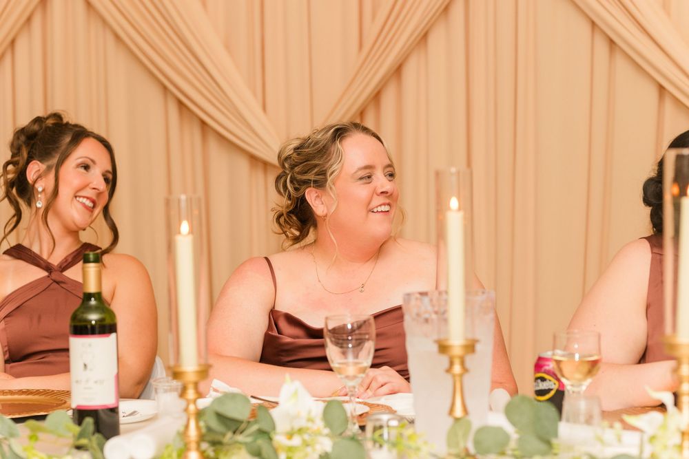 Bridesmaids in brown dresses laughing at a wedding reception table with candles and wine.