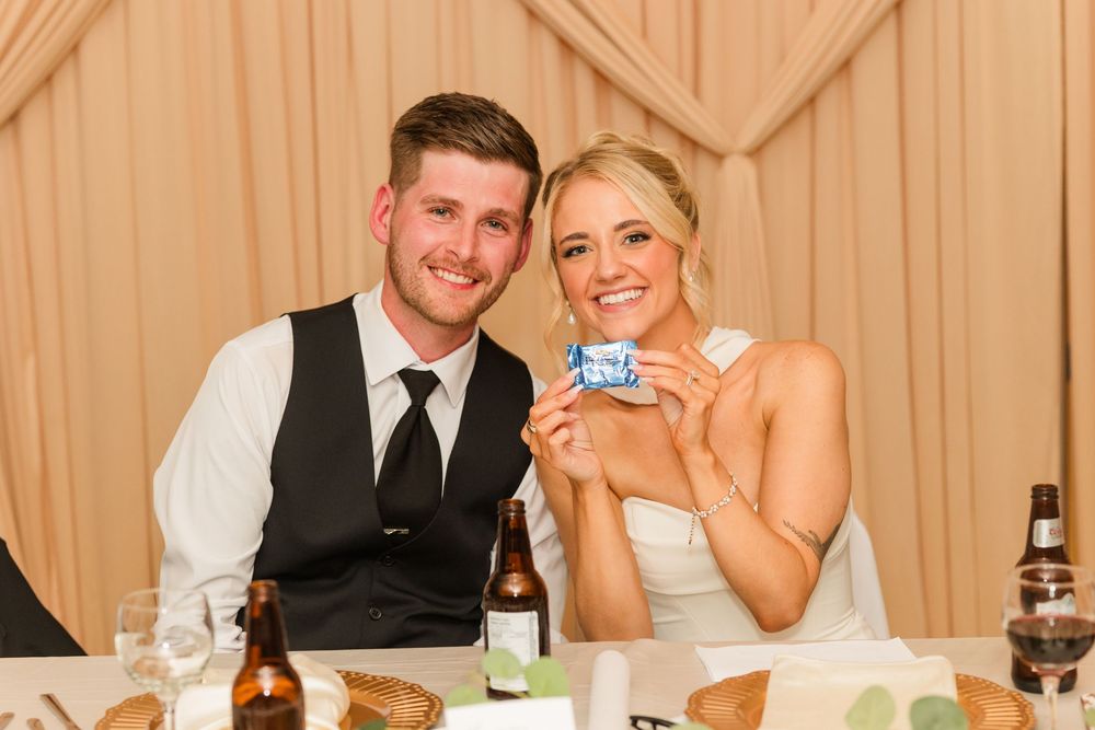 Smiling bride and groom sitting at wedding reception table, holding a small card, with beer bottles visible.
