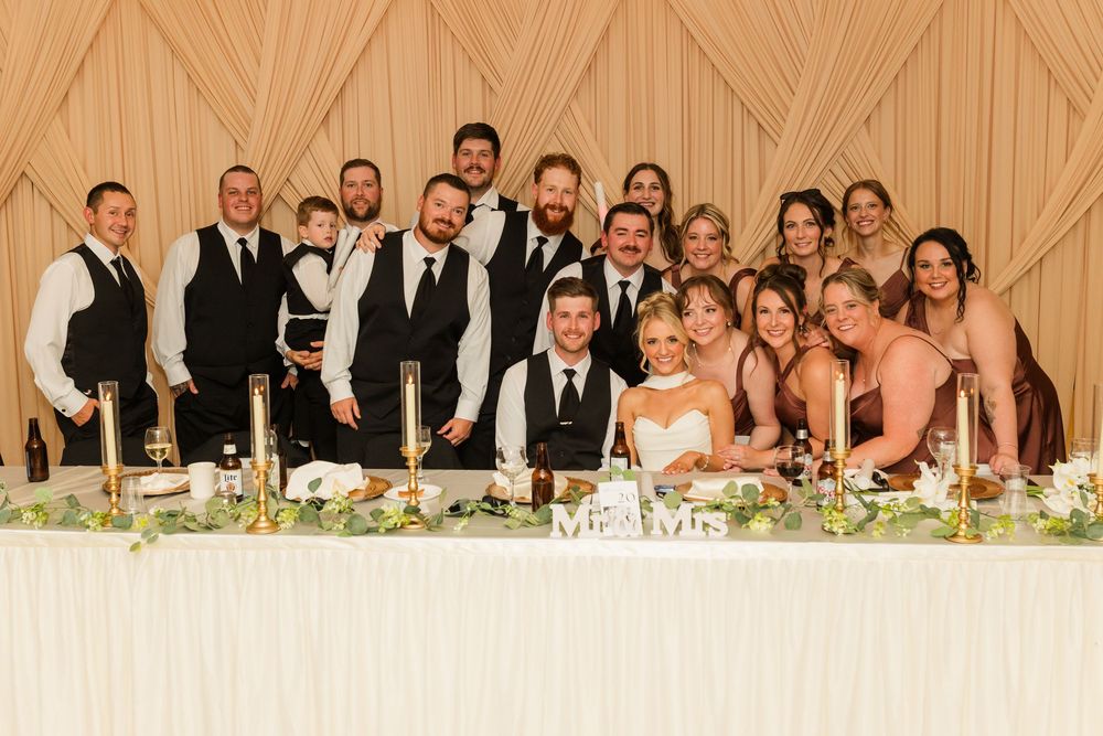 Wedding party poses together at elegantly decorated head table with greenery and candles at a formal reception.