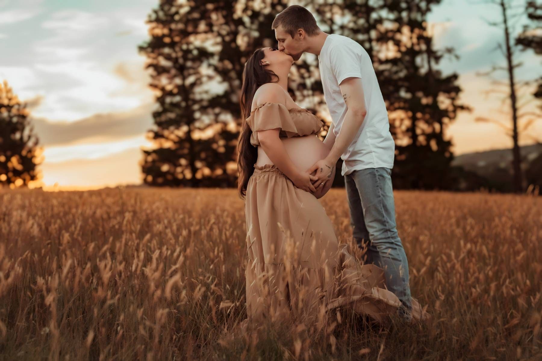 Pregnant couple sharing a romantic kiss in a golden field at sunset, with trees silhouetted in the background.