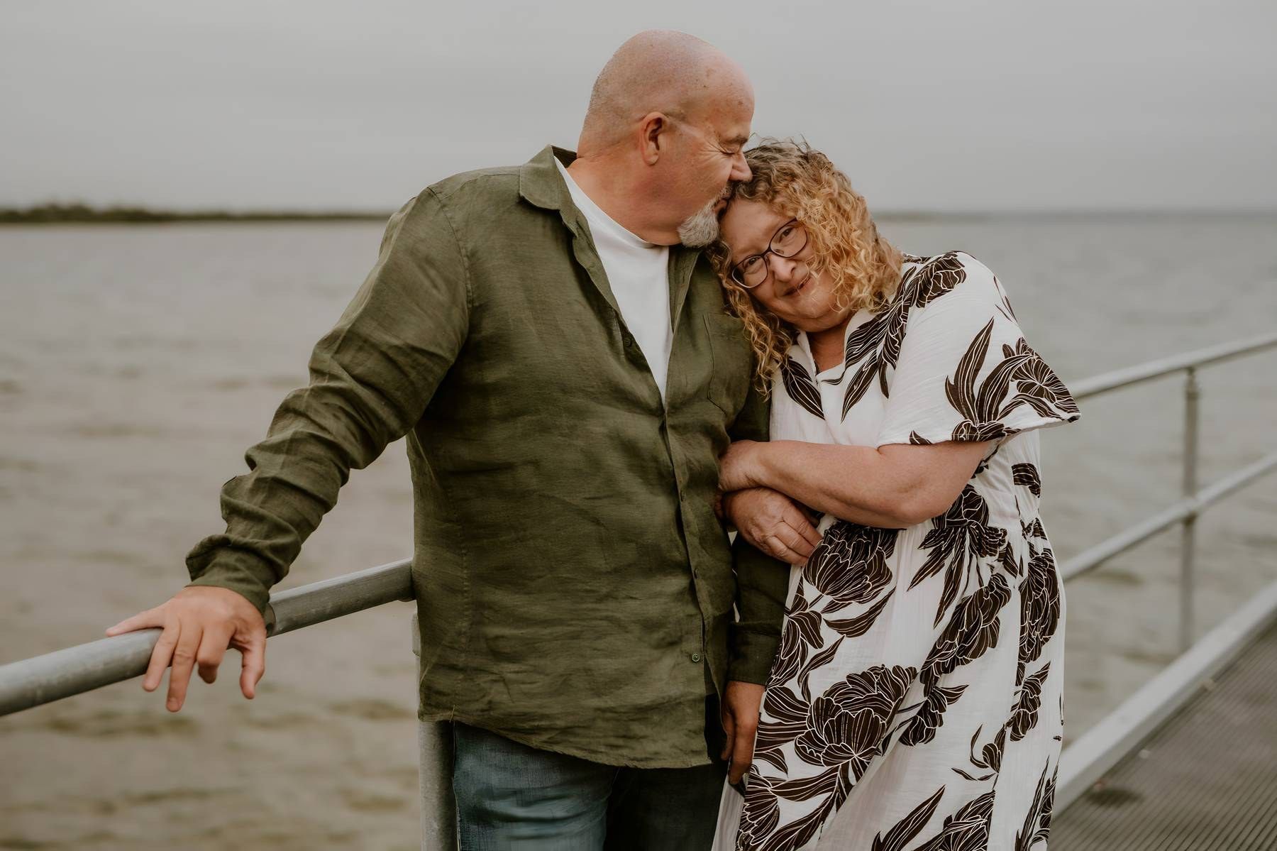 Couple embracing on a waterfront pier, man kissing woman's head, wearing casual attire on a cloudy day.