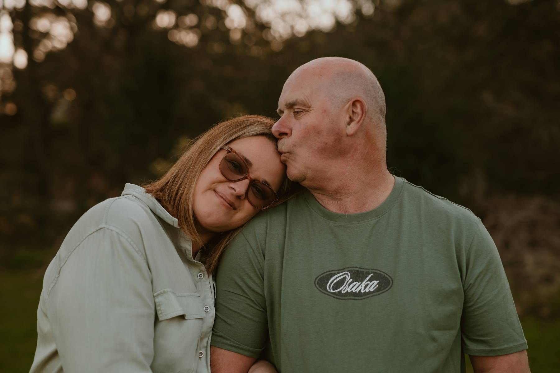 A bald man kisses a smiling woman wearing sunglasses outdoors at golden hour in a warm, romantic couple portrait.