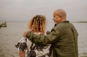 Couple embracing by the ocean, woman with curly hair in floral dress, bald man in green shirt, overcast sky.