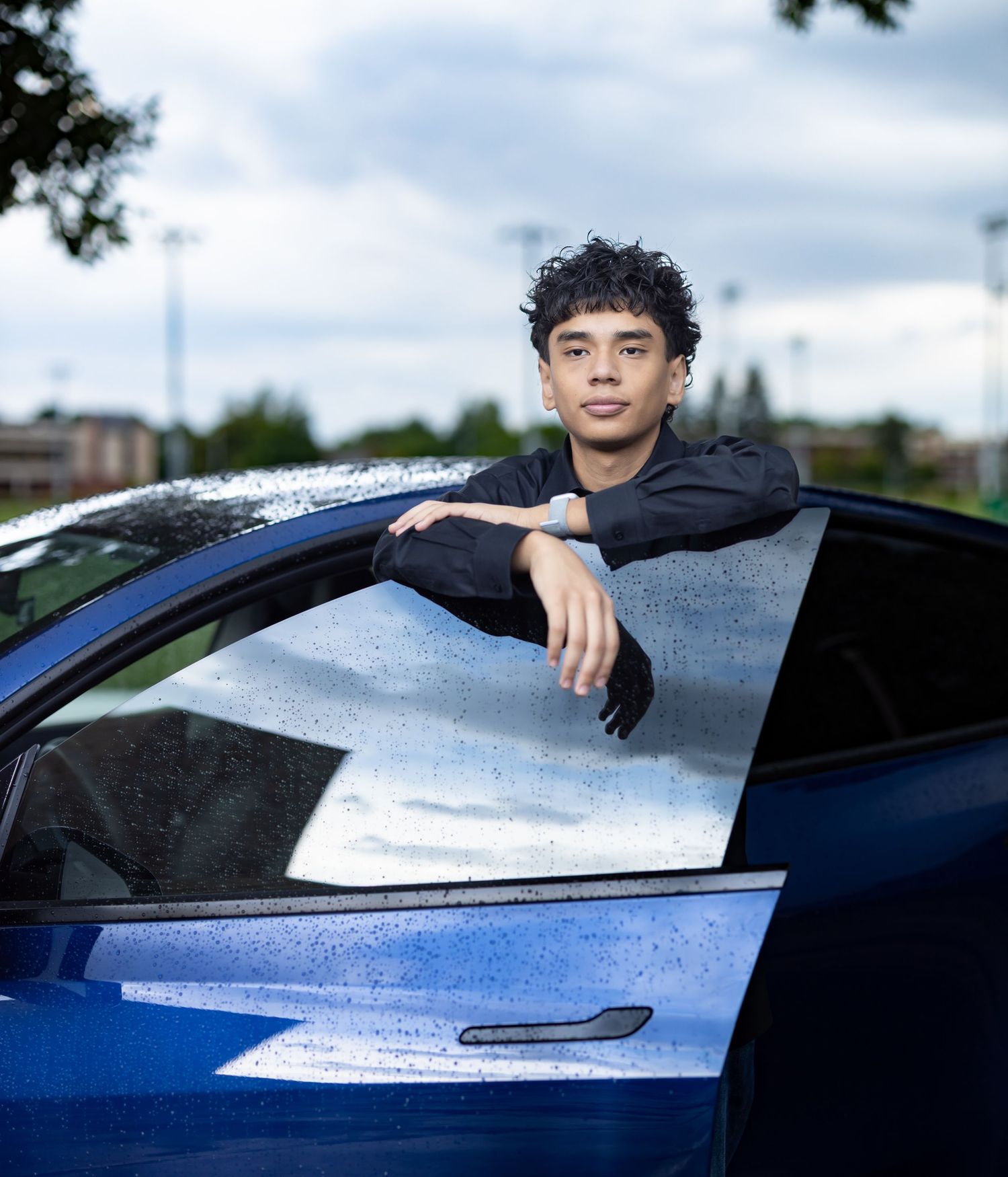 Young man in black outfit leaning out of blue car window on cloudy day.