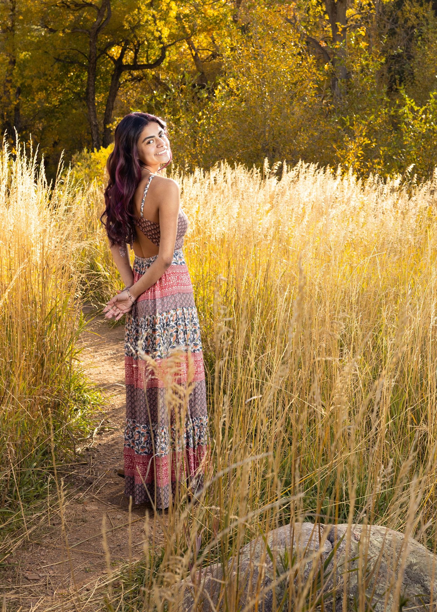 Young woman in colorful boho maxi dress standing on a path surrounded by golden tall grass and autumn trees.