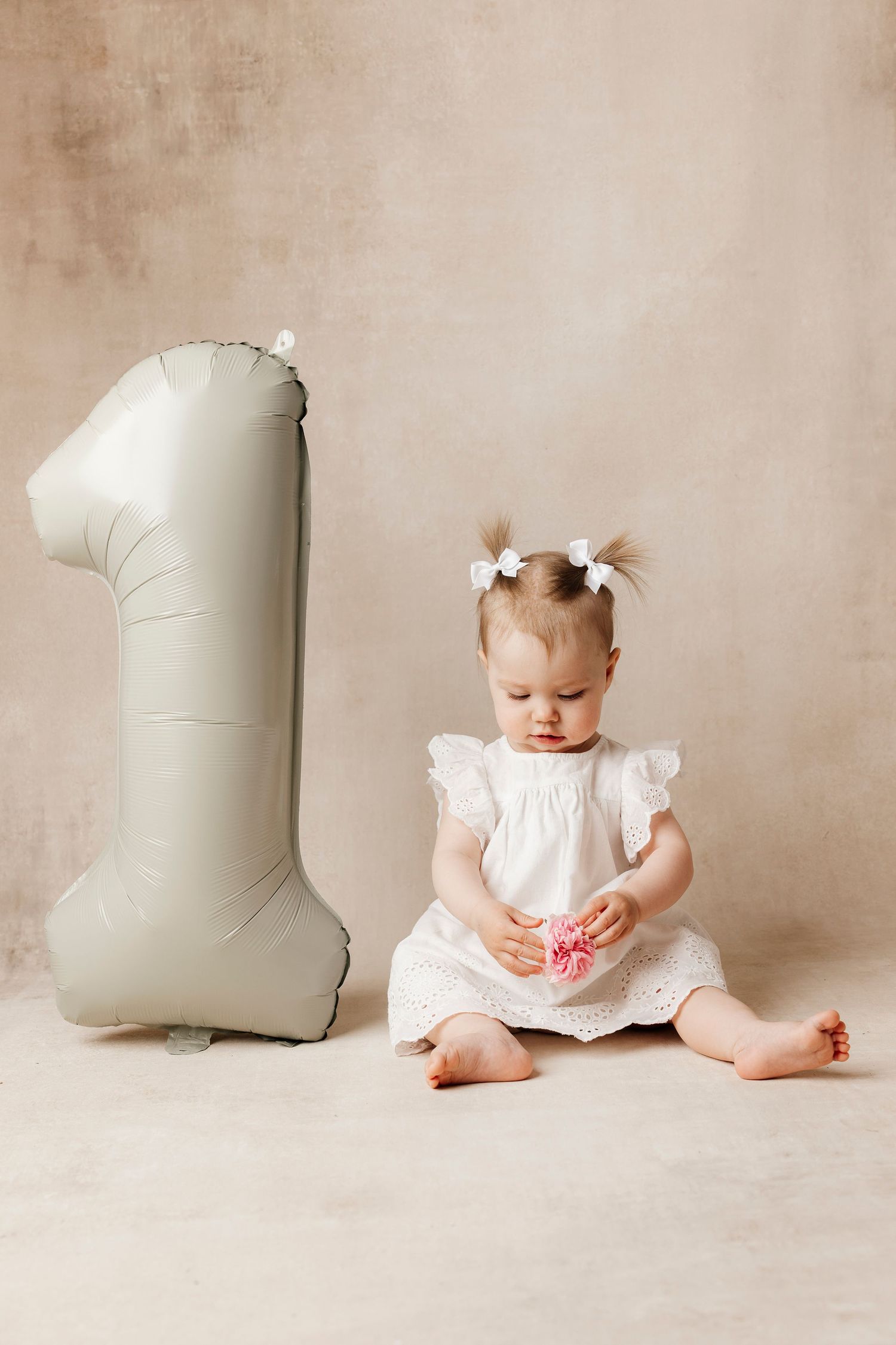 Cute baby girl in white dress sitting next to a number one balloon celebrating her first birthday.