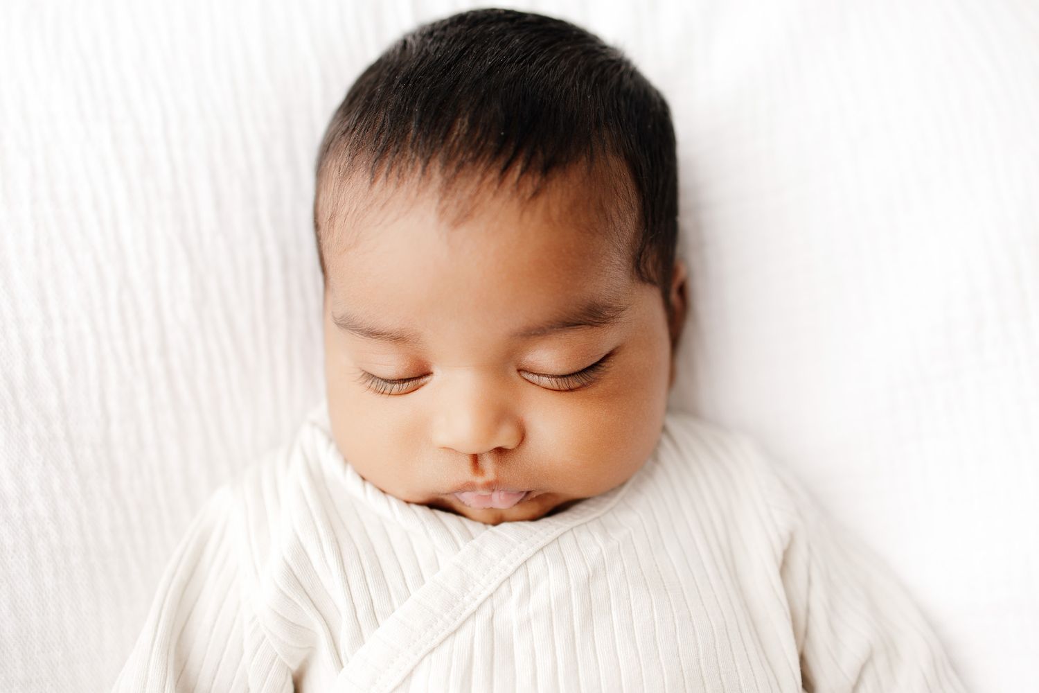 Peaceful newborn baby sleeping on white blanket, dark hair, soft skin, serene expression, professional newborn photography.