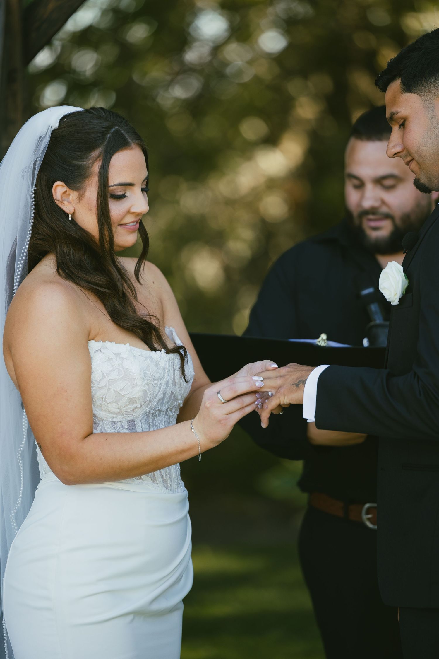 Bride smiles as she places wedding ring on groom's finger during outdoor ceremony, officiant standing behind them.