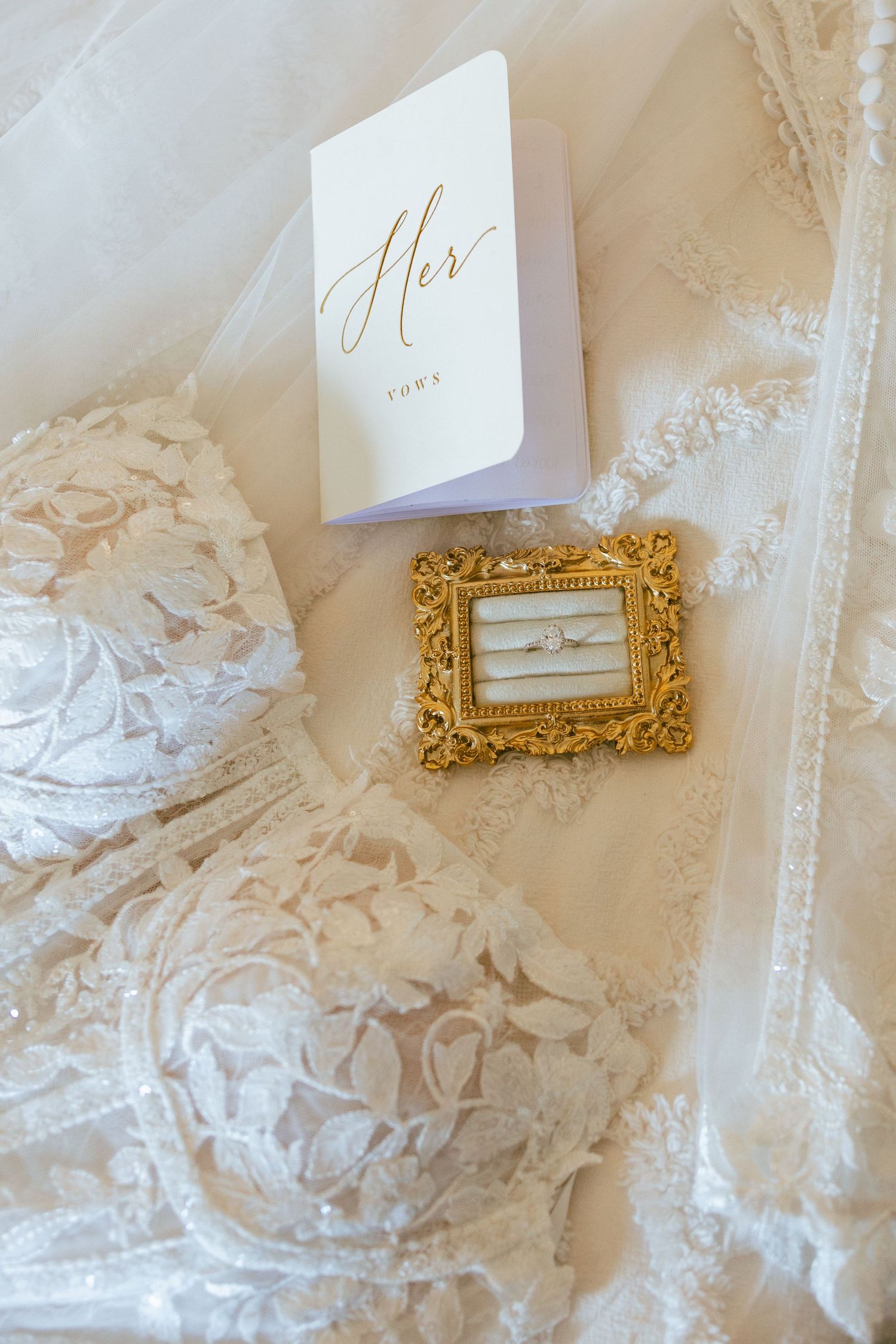 Bridal flat lay with white lace lingerie, gold ornate ring dish with diamond ring, and elegant Her vows card.