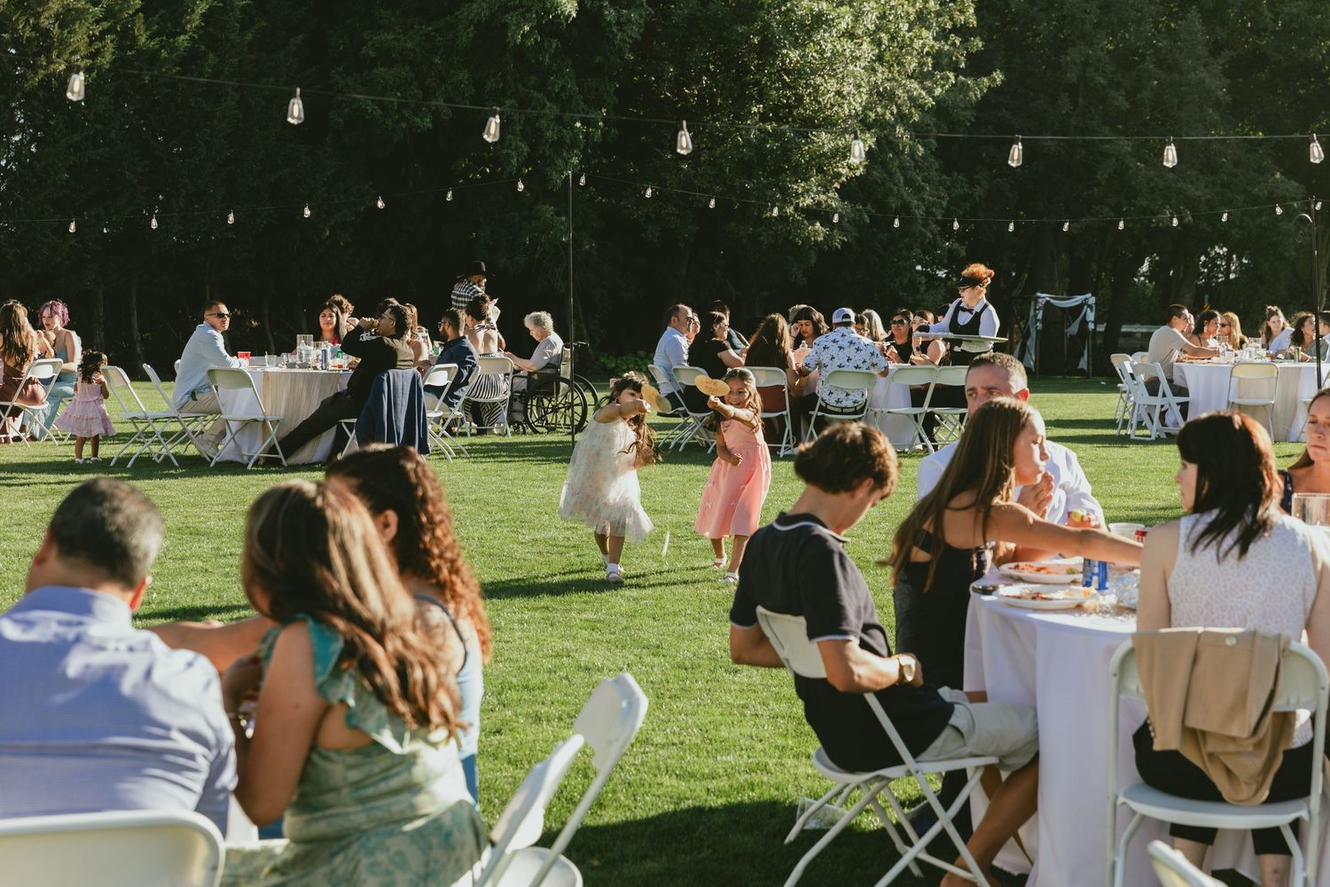 Outdoor wedding reception on green lawn with guests seated at white tables under string lights surrounded by trees.