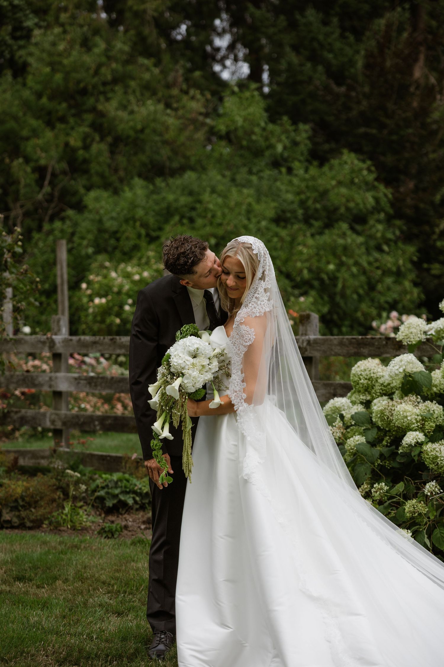 Bride in white lace gown and veil holds white bouquet as groom kisses her cheek in lush garden setting.