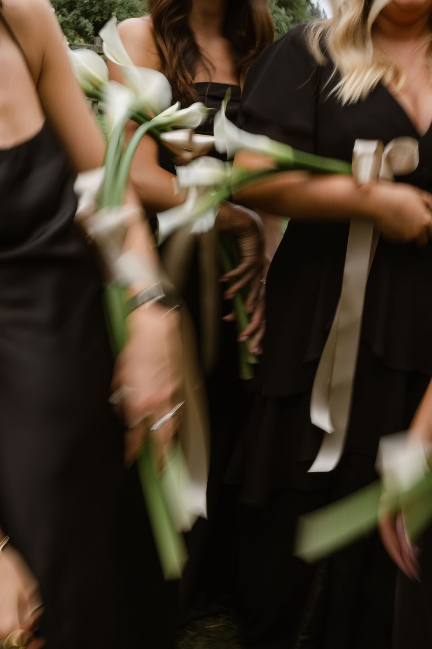Bridesmaids in black dresses holding white calla lily bouquets with ribbon, captured in artistic motion blur.