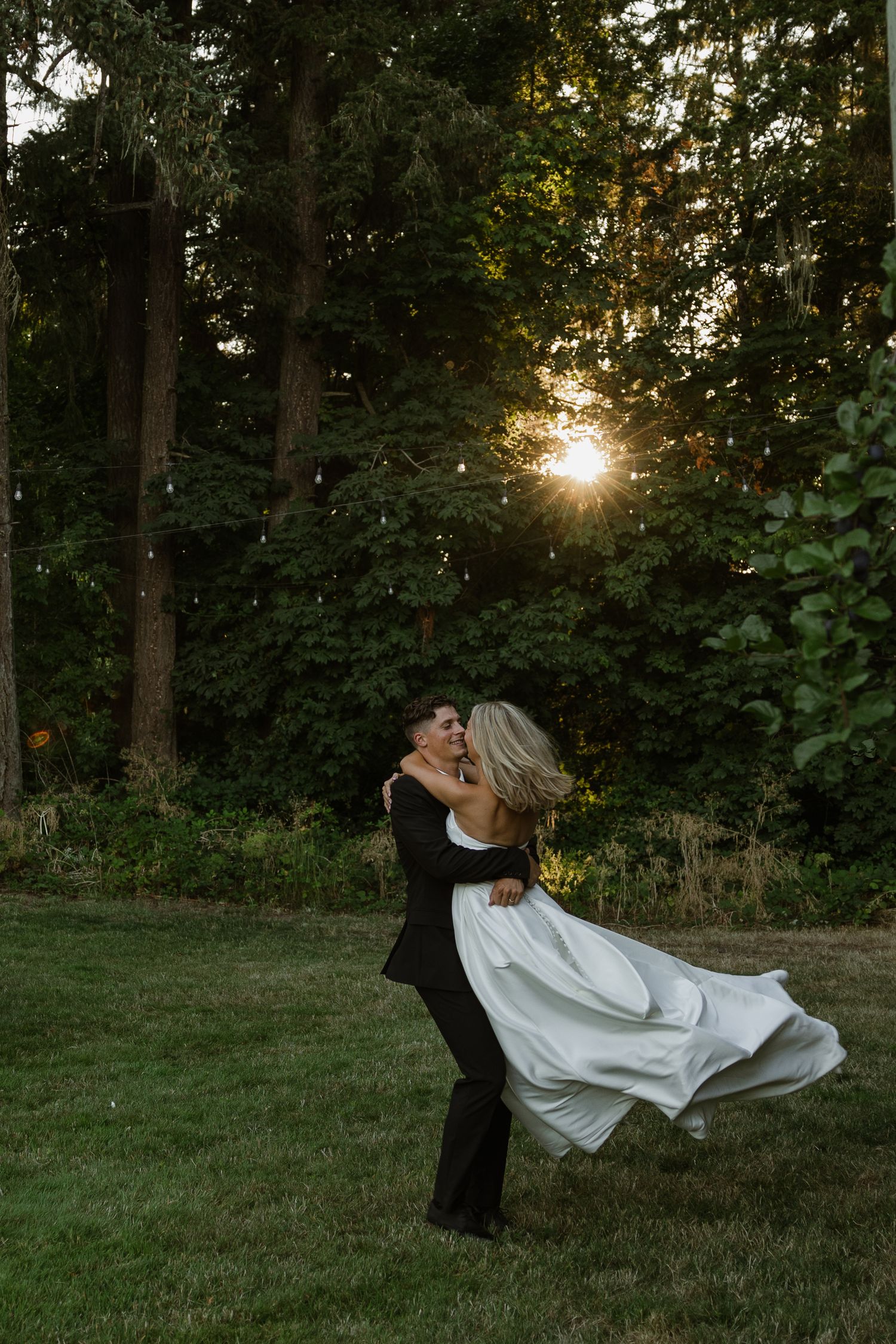 Groom lifting bride in white gown at sunset, surrounded by lush green trees in an outdoor wedding setting.
