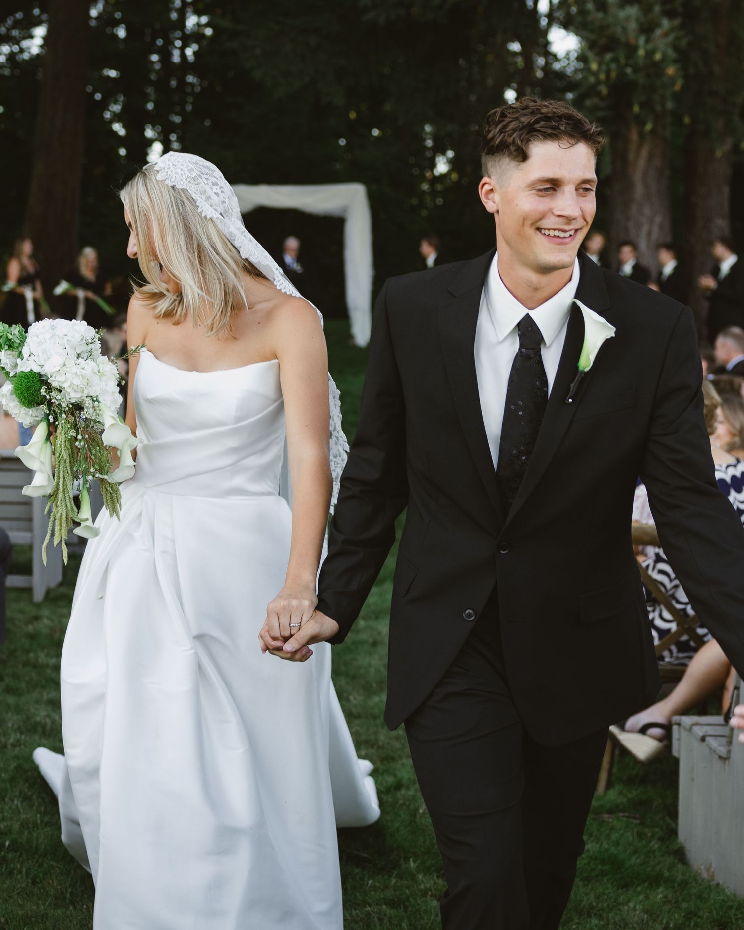 A smiling groom in a black suit holds hands with a bride in a white strapless gown and veil at an outdoor wedding.
