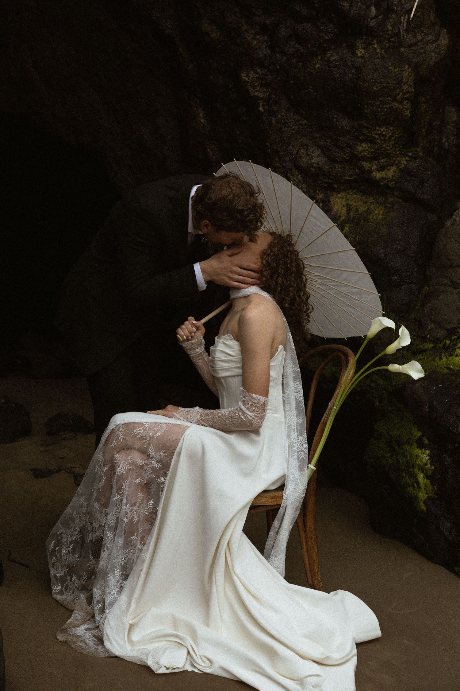 Bride in white lace gown holds parasol while kissing groom on sandy beach near dark rocky cliffs.