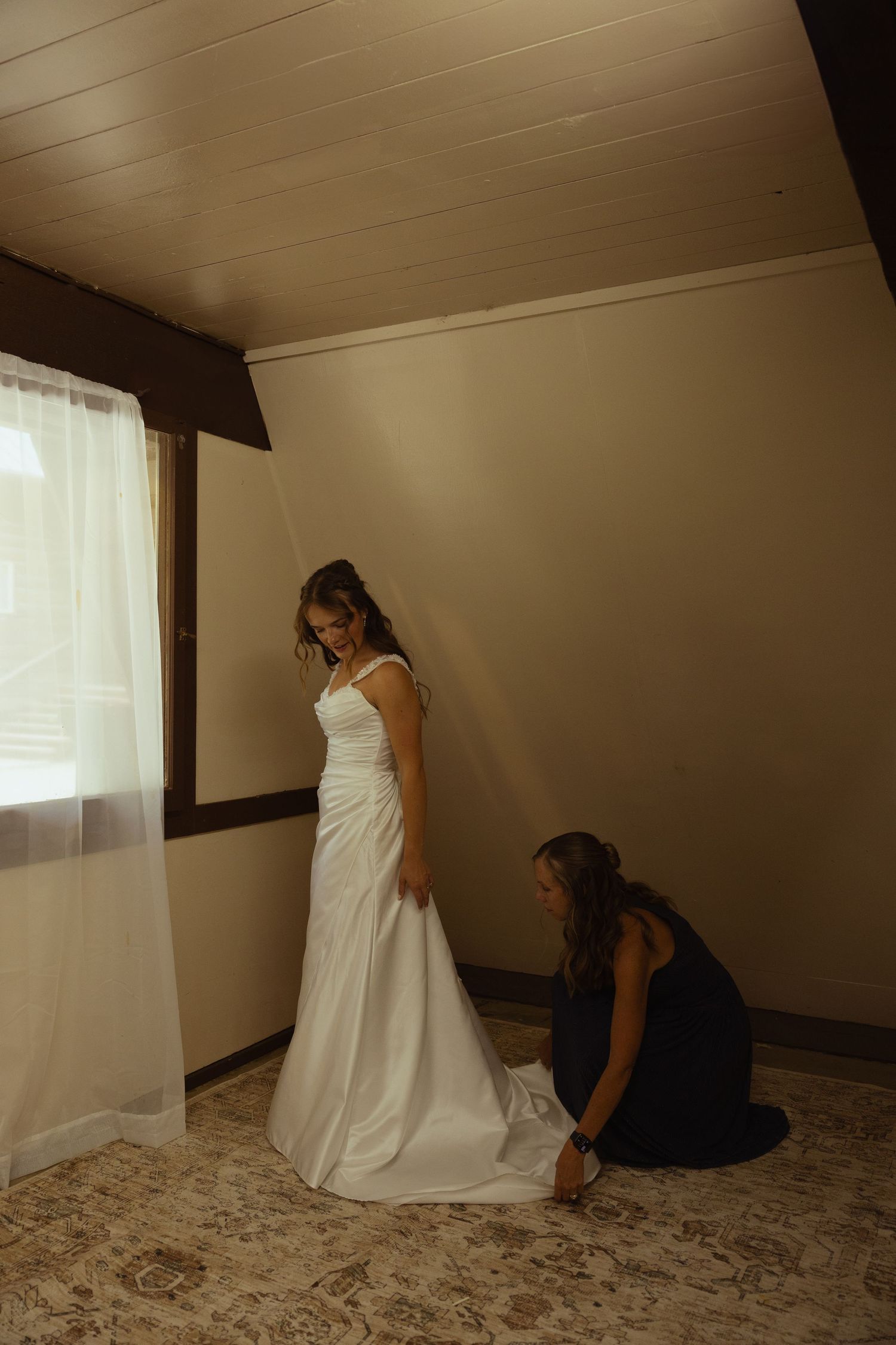 Bride in white wedding gown being helped by bridesmaid in navy dress adjusting her train in a softly lit room.