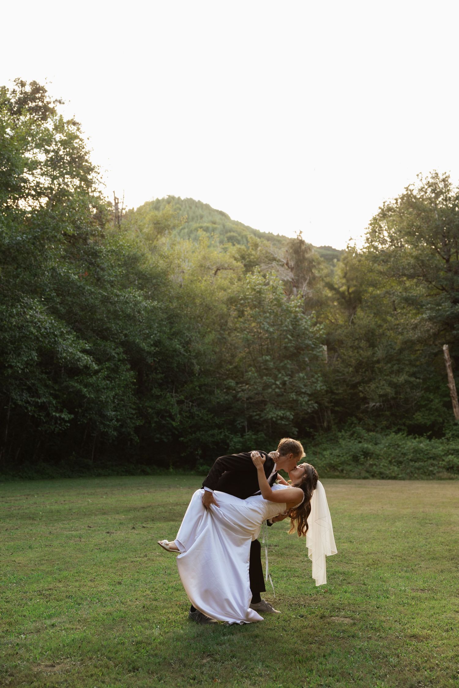 Groom lifts bride in white dress for a romantic kiss on a green lawn surrounded by lush trees and hills at sunset.
