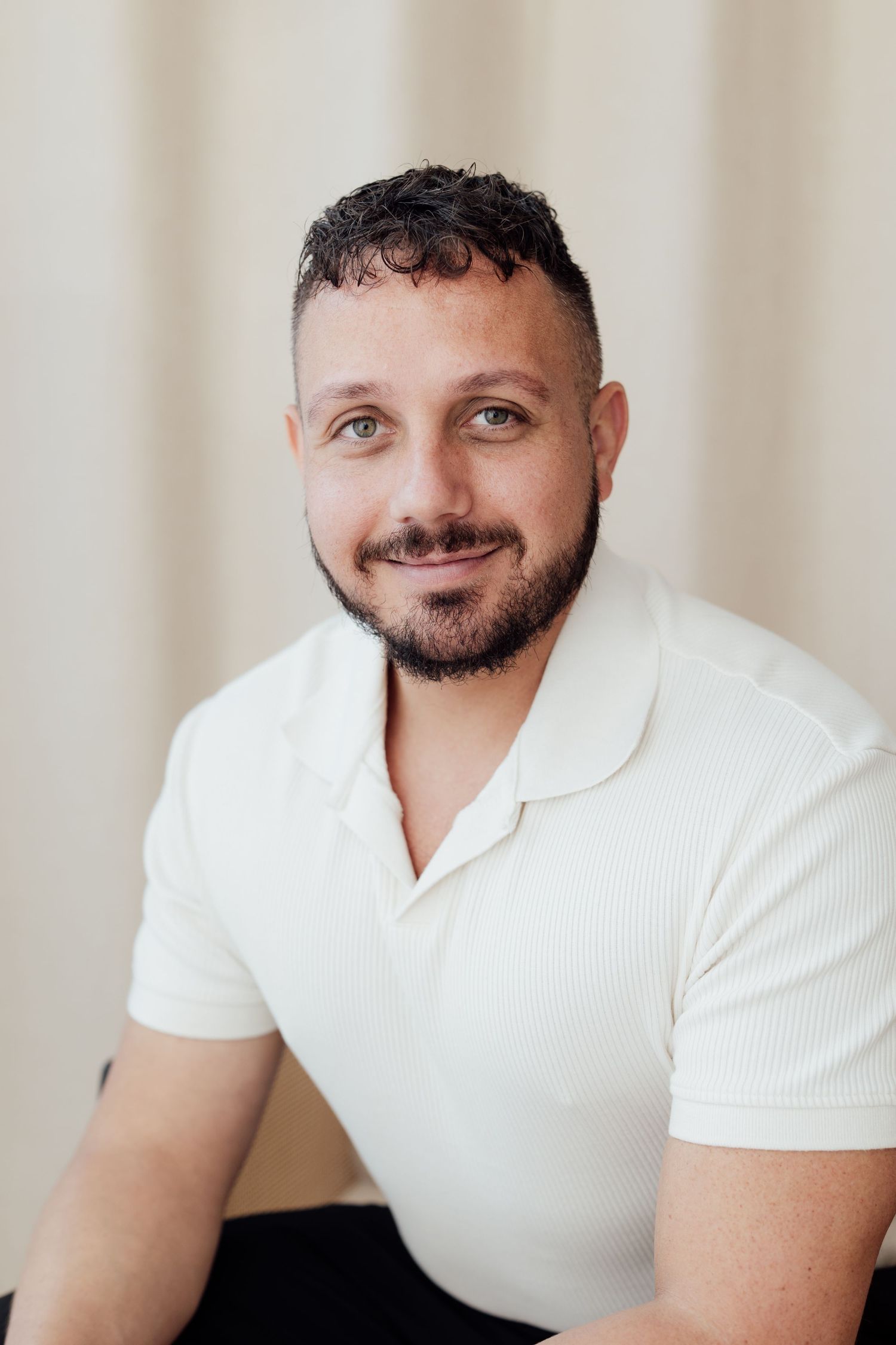 Smiling man with dark curly hair and beard wearing a white ribbed polo shirt, posed against a neutral beige background.
